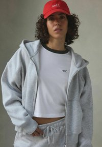 Šedá mikina na zip s předními kapsami, bílý crop top s vyšitým logem a červená čepice. Materiál je měkký, z bavlněné směsi; volný střih.