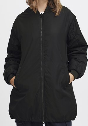 Personne portant une longue veste noire à capuche avec poignets élastiques et fermeture éclair frontale, les mains dans les poches latérales, vue du cou jusqu'au milieu des cuisses.