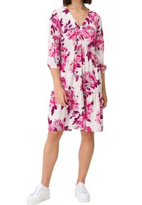 Femme portant une robe blanche jusqu'aux genoux avec des motifs floraux roses, des manches trois-quarts, et des baskets blanches à plateforme.