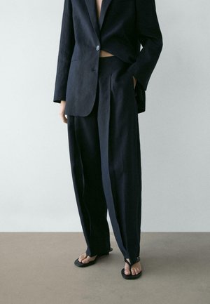 Persona che indossa un abito nero largo con una mano in tasca e sandali infradito neri, in piedi su un pavimento semplice contro una parete chiara.