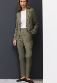 Olivzaļš pielāgots uzvalks ar izgriezuma atlokiem, sānu kabatām un atbilstošām biksēm. Balts T-krekls ar apaļu kaklu zemūdens. Brūnas slip-on kurpes.