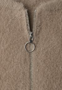 Veste camel à fermeture éclair en matière douce et duveteuse. Elle est dotée d'une fermeture éclair argentée avec un tirette ronde et d'un col légèrement côtelé.