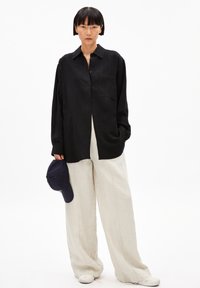 Schwarze Hemdjacke mit einer Tasche, kombiniert mit hellbeigen weiten Hosen. Das Model hält eine dunkelblaue Mütze und steht vor einem weißen Hintergrund.