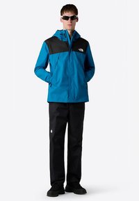 Blue waterproof jacket with black accents, zippered front, elastic cuffs, paired with black pants and black shoes. Model wearing sunglasses.