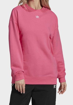 Femme portant un sweat-shirt Adidas rose et un pantalon Adidas noir, debout devant un fond clair uni.