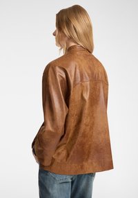 Giubbotto di pelle marrone con una texture liscia e invecchiata, caratterizzato da una vestibilità morbida e un colletto rotondo. Cuciture visibili lungo la cucitura posteriore.