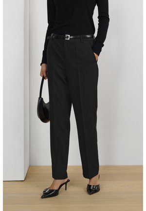 Vrouw draagt zwarte plissé broek met hoge taille, zwarte riem, zwarte puntige hakken met gesp, zwart shirt met lange mouwen en houdt een kleine zwarte handtas vast.
