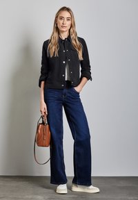 Svart jacka med tryckknappar, vit topp, blå vida jeans och beige skor. Håller en brun bucket bag med en rem.