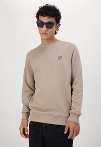 Beige pulóver hosszú ujjakkal, kerek nyakkivágással, bordázott mandzsettákkal és alsó szegéllyel; a mellkason egy kis, sárga hímzett logó található.