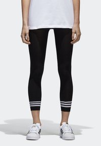 Černé legíny s žebrovanou strukturou a bílými vodorovnými pruhy na lemování. V kombinaci s bílými sportovními botami a volnou bílou košilí.