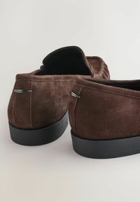 Bruine suède instappers met een zwarte rubberen zool, voorzien van subtiele stiksels en metalen accenten op de hiel. Tijdloos ontwerp, ronde vorm.