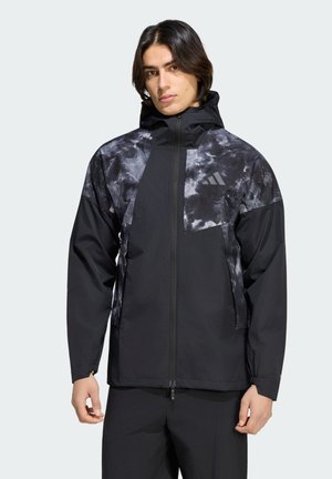 Persona joven con cabello oscuro de media longitud lleva una chaqueta de patrón tie-dye en negro y gris, con cremallera frontal y capucha.