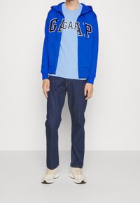 Sweat à capuche zippé bleu avec inscription blanche, t-shirt bleu clair en dessous, jean foncé et baskets beiges. Ensemble de vêtements décontractés.