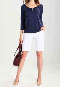 Haut à manches longues marine avec encolure henley et trois boutons, accompagné de shorts blancs. Accessoirisé avec un sac à main bordeaux et des talons nus.