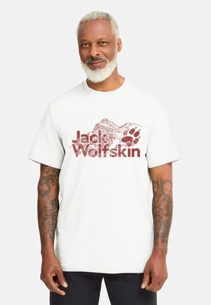 Uomo con barba grigia che indossa una maglietta bianca a maniche corte con grafica di montagna e testo "Jack Wolfskin", in piedi davanti a uno sfondo chiaro uniforme.