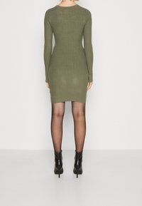 Robe à manches longues côtelée vert olive avec une silhouette ajustée. Une paire de bottines noires à talons hauts est visible sous l'ourlet qui arrive au-dessous du genou.