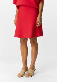 Gonna A-line rossa realizzata in tessuto leggero, abbinata a sandali beige slip-on con un morbido design incrociato e punta aperta.