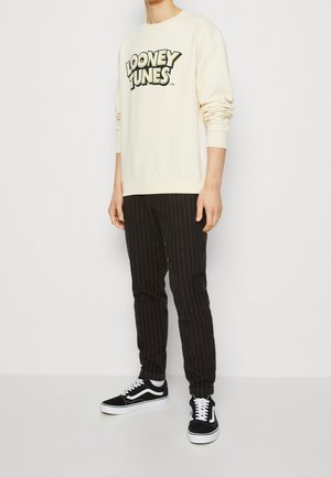 Cremefarbener Sweatshirt mit "Looney Tunes"-Logo, schwarze gestreifte Hose und schwarze Vans-Sneaker. Lässiges Outfit mit lockerer Passform und strukturiertem Stoff.
