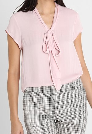 Femme portant un chemisier rose clair à manches courtes avec un nœud à l'avant du col, associé à un pantalon à carreaux noir et blanc.