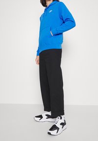 Osoba oblečená v jasně modré mikině Nike, černých kalhotách a černobílých teniskách stojí na bílém pozadí.