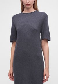 Femme portant une robe-pull courte à manches courtes gris foncé unie, debout devant un fond blanc.