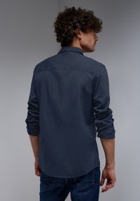 Camisa de botones azul marino con cuello, mangas enrolladas y textura suave; diseño ajustado con dobladillo recto y detalle en el yugo trasero.
