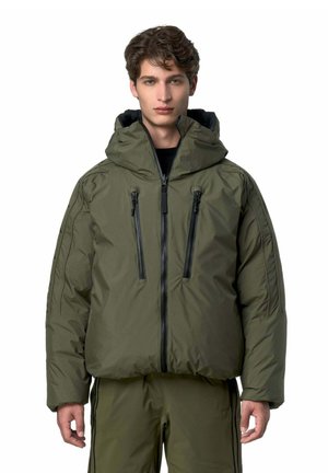 Jeune homme portant une veste matelassée vert olive avec une capuche et un pantalon assorti, debout sur fond blanc.