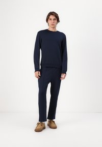 Navyblauwe sweatshirt met ronde hals en lange mouwen, gecombineerd met bijpassende tapered broek. Model draagt tan suède schoenen met gestructureerde zolen.