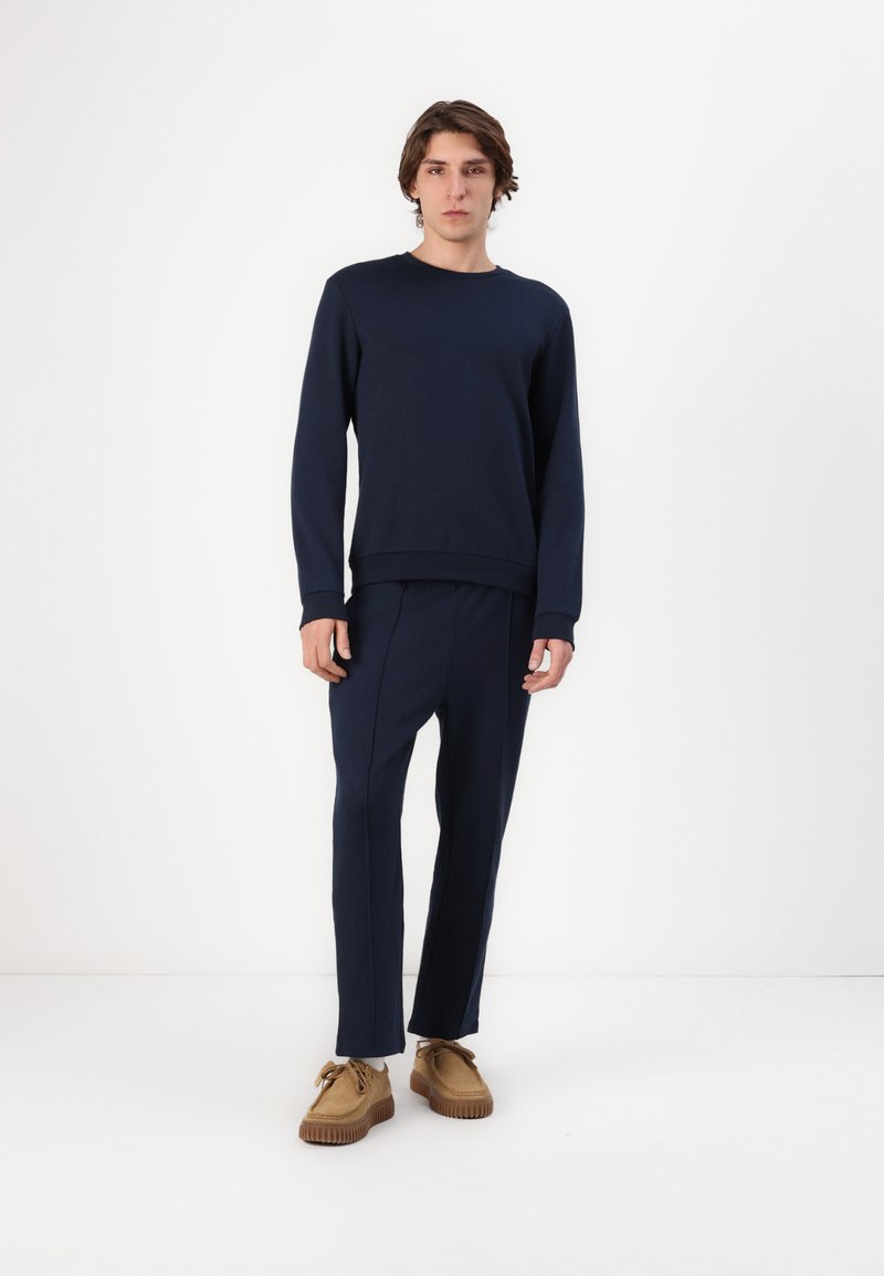 Navyblauwe sweatshirt met ronde hals en lange mouwen, gecombineerd met bijpassende tapered broek. Model draagt tan suède schoenen met gestructureerde zolen.