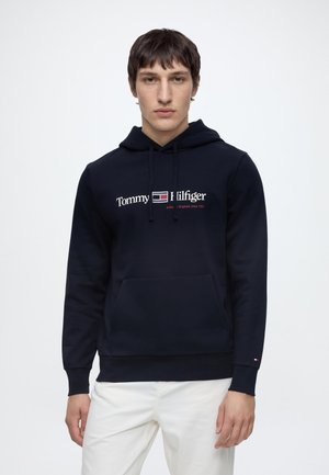 Jeune homme portant un sweat à capuche bleu marine Tommy Hilfiger avec un pantalon blanc sur un fond clair uni.