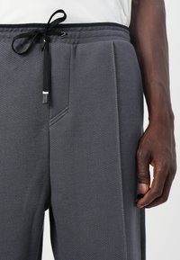 Pantaloni de jogger gri, dintr-un material texturat cu o suprafață netedă, având un tiv elastic și un șnur negru cu vârf argintiu.