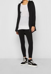 En ung kvinna står klädd i en svart zip-up hoodie, en vit stickad topp, svarta leggings och svarta sneakers med vita detaljer framför en grå bakgrund.