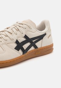 Beige zamšas sporta apavi ar melniem ādas svītrām, apaļa purngala forma, plakanās auklas un gumijas zole, kas apvieno zelta "asics" zīmolu sānos.
