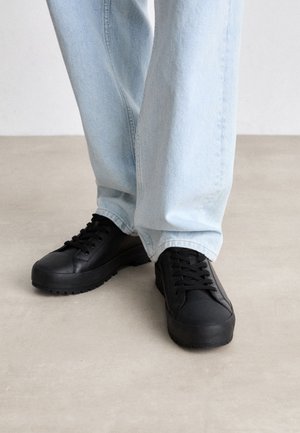 Osoba ubrana w jasnoniebieskie dżinsy i czarne sznurowane buty na platformie stojąca na jasnoszarej podłodze.