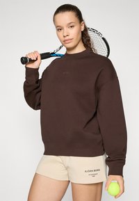 Sweat-shirt marron avec un col côtelé et un logo, associé à des shorts beiges arborant un texte. Le modèle tient une raquette de tennis et une balle.