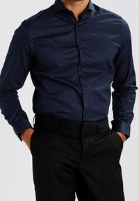 Námořnicky modrá košile na knoflíky s texturovaným vzorem, dlouhými rukávy a klasickým límcem. V kombinaci s černými kalhotami.