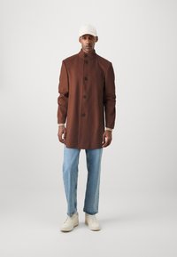 Bruin wollen mix jas met een hoge kraag, knoopsluiting aan de voorkant en zijzakken, gedragen met lichtblauwe jeans en witte sneakers.