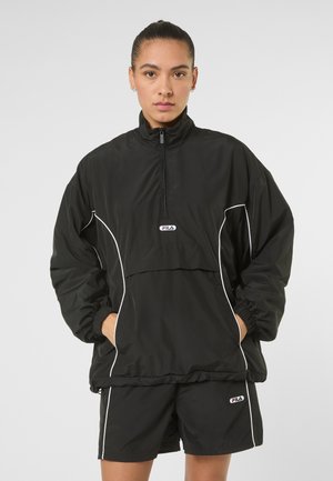 Kvinde med håret bundet tilbage iført sort FILA-jakke med hvide pipingdetaljer og matchende shorts, hænderne i jakkelommerne, neutral udtryk.