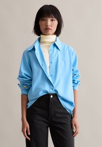 Light blue button-up shirt with a glossy texture, oversized fit, and rolled sleeves, paired with high-waisted black jeans.