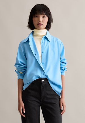 Hellblaues Button-Up-Hemd mit glatter Textur, oversized Passform und hochgerollten Ärmeln, kombiniert mit hoch taillierten schwarzen Jeans.