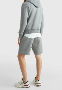 Felpa grigia con tasca a marsupio, abbinata a pantaloni corti grigi coordinati. Camicia bianca sotto. Sneakers con una combinazione di dettagli bianchi e blu navy.