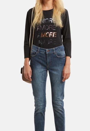 Vrouw draagt een zwart shirt met lange mouwen met glinsterende tekst "AMORE" en "MORE", blauwe jeans en een kleine bruine schoudertas.