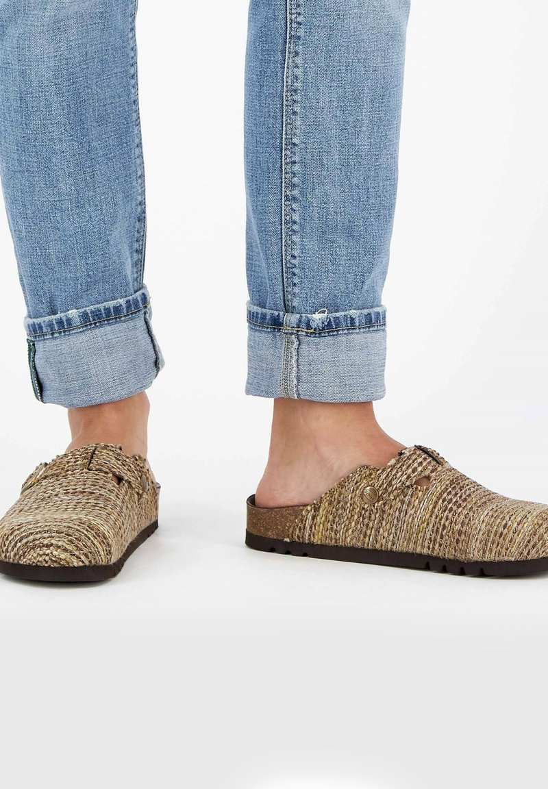 Sandales brunes tissées avec une surface texturée, dotées d'un design sans arrière, d'une sangle ajustable et d'une semelle épaisse. Associées à des jeans bleus retroussés.