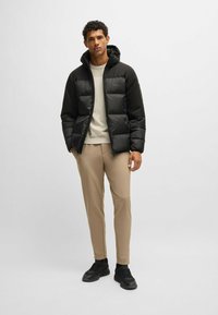 Black puffer jacket with hood, featuring a quilted design and logo on the chest. Worn over a light beige sweater and beige tapered pants.