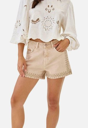 Mujer con pantalones cortos bordados beige y una blusa blanca de manga larga con patrones florales recortados, mano en el bolsillo sobre un fondo blanco.