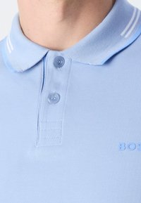 Homme portant un polo bleu clair avec deux boutons fermés et un col côtelé avec des rayures blanches.