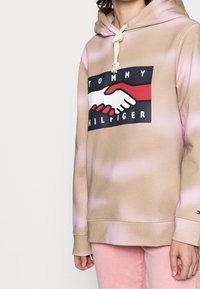 Beige mikina s kapucí, s pink gradientovým barvením, na které je výrazný grafický prvek zobrazení rukou, které si podávají ruku, a text "TOMMY HILFIGER". Kapuce se stahovací šňůrkou, měkká textura.