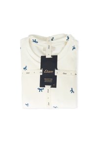 Camiseta de color crema con bordado de lazo azul marino, doblada de manera ordenada, con una etiqueta que muestra el nombre de la marca y la información de talla.