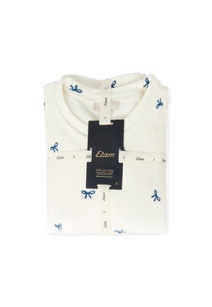 Krämfärgad T-shirt med mörkblå bågbroderi, vikt snyggt, med en etikett som visar varumärke och storleksinformation.