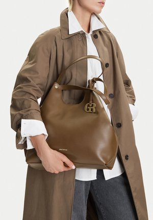 Mujer con un abrigo trench marrón y camisa blanca sosteniendo un gran bolso de cuero marrón con un charm y logo dorados con las letras "GR".